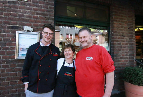 Drei lächelnde Restaurantmitarbeiter vor einem Ziegelstein-Gebäude stehen neben einem Fenster mit Menütafel.